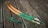 Two curved wooden objects with green and brown hues rest on a wooden plank surface. The items appear to be archery bow limbs with intricate patterns. A coiled green string with fluffy sections is positioned nearby.