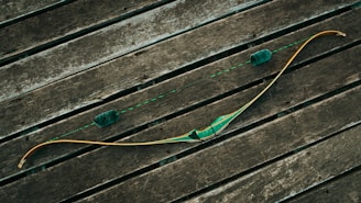 A traditional wooden recurve bow rests on a rustic wooden surface, featuring a green grip and string dampeners. The bowstring is taut and aligned symmetrically with the limbs, while the wood planks create a textured backdrop.