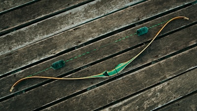 A traditional wooden recurve bow rests on a rustic wooden surface, featuring a green grip and string dampeners. The bowstring is taut and aligned symmetrically with the limbs, while the wood planks create a textured backdrop.