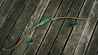 A traditional bow with a green grip and accents lies across weathered wooden planks. The bowstring is taut, featuring two fuzzy silencers. The wood of the bow has a natural finish, highlighting its craftsmanship.