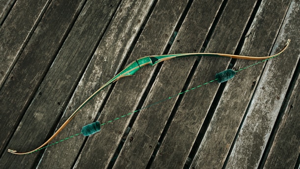A traditional bow with a green grip and accents lies across weathered wooden planks. The bowstring is taut, featuring two fuzzy silencers. The wood of the bow has a natural finish, highlighting its craftsmanship.