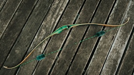 A traditional bow with a green grip and accents lies across weathered wooden planks. The bowstring is taut, featuring two fuzzy silencers. The wood of the bow has a natural finish, highlighting its craftsmanship.