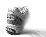 A close-up view of a sports shoe with a focus on its detailed sole and stitching. The shoe features a prominent logo and intricate patterns on its surface, set against a blurred background that highlights its design.