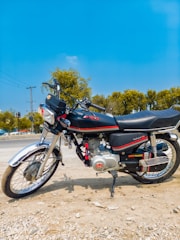 A motorcycle with a sleek design is parked on a dirt surface. The bike has a black and silver color scheme with red accents and is positioned against the backdrop of a clear blue sky and green trees. It features a detailed engine and shiny chrome parts reflecting sunlight.
