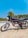 A motorcycle with a sleek design is parked on a dirt surface. The bike has a black and silver color scheme with red accents and is positioned against the backdrop of a clear blue sky and green trees. It features a detailed engine and shiny chrome parts reflecting sunlight.