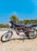 A motorcycle with a sleek design is parked on a dirt surface. The bike has a black and silver color scheme with red accents and is positioned against the backdrop of a clear blue sky and green trees. It features a detailed engine and shiny chrome parts reflecting sunlight.