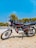 A motorcycle with a sleek design is parked on a dirt surface. The bike has a black and silver color scheme with red accents and is positioned against the backdrop of a clear blue sky and green trees. It features a detailed engine and shiny chrome parts reflecting sunlight.