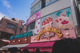 A vibrant street scene featuring colorful storefronts with neon signage. One sign depicts a female figure in a pose, promoting a burlesque establishment. Adjacent is a sign for Santa Monica Crepes, adorned with illustrations of crepes topped with strawberries and whipped cream. The building surfaces display a mix of bricks and tiles, with visible polka dot patterns. A clear blue sky enhances the bright and lively atmosphere.