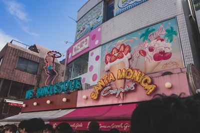 A vibrant street scene featuring colorful storefronts with neon signage. One sign depicts a female figure in a pose, promoting a burlesque establishment. Adjacent is a sign for Santa Monica Crepes, adorned with illustrations of crepes topped with strawberries and whipped cream. The building surfaces display a mix of bricks and tiles, with visible polka dot patterns. A clear blue sky enhances the bright and lively atmosphere.