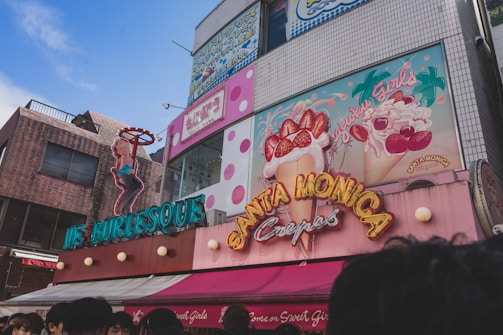 A vibrant street scene featuring colorful storefronts with neon signage. One sign depicts a female figure in a pose, promoting a burlesque establishment. Adjacent is a sign for Santa Monica Crepes, adorned with illustrations of crepes topped with strawberries and whipped cream. The building surfaces display a mix of bricks and tiles, with visible polka dot patterns. A clear blue sky enhances the bright and lively atmosphere.