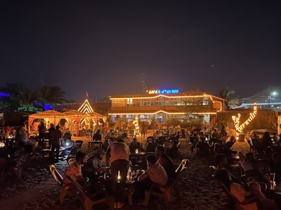 A vibrant beachside bar is bustling with people at night, featuring warm lighting and lively decor. Tables and chairs are arranged on the sand, with patrons enjoying the atmosphere under a clear, starry sky. The bar is adorned with decorative lights, adding a festive ambiance.