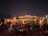 A vibrant beachside bar is bustling with people at night, featuring warm lighting and lively decor. Tables and chairs are arranged on the sand, with patrons enjoying the atmosphere under a clear, starry sky. The bar is adorned with decorative lights, adding a festive ambiance.