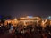 A vibrant beachside bar is bustling with people at night, featuring warm lighting and lively decor. Tables and chairs are arranged on the sand, with patrons enjoying the atmosphere under a clear, starry sky. The bar is adorned with decorative lights, adding a festive ambiance.