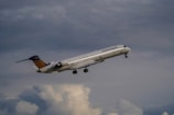 A commercial airplane with the Lufthansa Regional logo is taking off or climbing through a cloudy sky. The aircraft's design includes a sleek fuselage and multiple windows, with landing gear extended. The tail section features a distinctive blue pattern with orange dots.