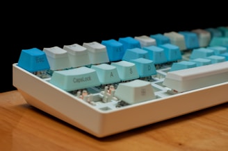 A close-up view of a mechanical keyboard featuring keycaps in shades of blue and white. The keyboard is placed on a wooden surface and has visible mechanical switches underneath the keycaps.