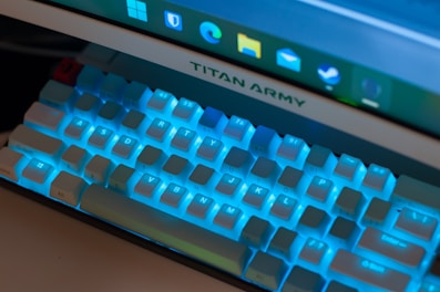 A close-up view of a mechanical keyboard with blue backlighting beneath the keys, positioned in front of a monitor displaying a taskbar with various application icons. The monitor has the brand name 'TITAN ARMY' visible.