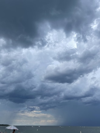 Dark, heavy clouds dominate the sky, indicating an approaching storm over a large body of water. The horizon is visible, with a person holding an umbrella on the beach in the bottom left corner of the image. A few boats can be seen in the distance on the water.