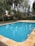 A serene outdoor swimming pool with clear blue water is surrounded by a stone deck. Tall trees with lush green foliage provide a natural backdrop, creating a peaceful and inviting environment. In the distance, a wooden fence and a hint of a building with a terracotta roof can be seen.