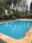 A serene outdoor swimming pool with clear blue water is surrounded by a stone deck. Tall trees with lush green foliage provide a natural backdrop, creating a peaceful and inviting environment. In the distance, a wooden fence and a hint of a building with a terracotta roof can be seen.