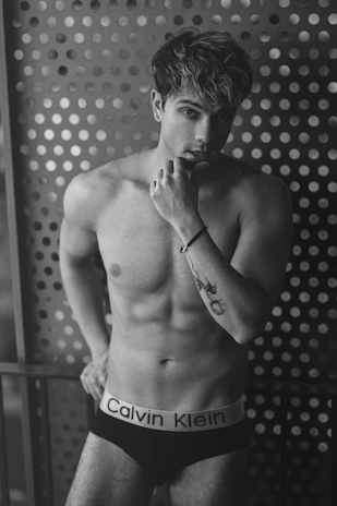 A shirtless man with a muscular build is posing with his left hand resting against his chin. He is wearing black Calvin Klein underwear and has a tattoo on his left forearm. The background consists of a perforated metallic wall with circular holes.