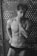 A shirtless man with a muscular build is posing with his left hand resting against his chin. He is wearing black Calvin Klein underwear and has a tattoo on his left forearm. The background consists of a perforated metallic wall with circular holes.