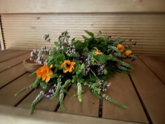 A bundle of fresh flowers and herbs is arranged on a wooden surface. Bright orange flowers stand out against a backdrop of green leaves and small purple blossoms, creating a natural and rustic aesthetic. The setting appears indoors, and the warm lighting adds a cozy touch.