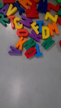 Colorful plastic alphabet letters scattered across a light gray surface. The letters are vibrant, including red, blue, yellow, green, orange, and purple shades. They appear to be jumbled and are likely used for educational or playful purposes.