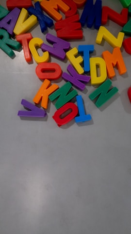 Colorful plastic alphabet letters scattered across a light gray surface. The letters are vibrant, including red, blue, yellow, green, orange, and purple shades. They appear to be jumbled and are likely used for educational or playful purposes.