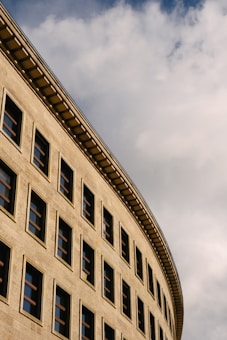A curved, modern building facade with rows of square windows set against a sky filled with fluffy clouds.