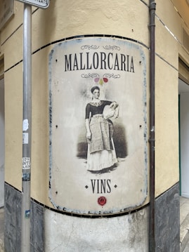 A vintage-style sign is mounted on a corner wall, depicting a woman in traditional attire holding a container. The text on the sign reads 'Mallorcaria' at the top and 'Vins' at the bottom. The sign appears weathered, adding an old-world charm.