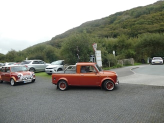 A vintage orange car is parked on a paved area with other vehicles nearby, set against a backdrop of lush green hills and trees. There are informational signposts and a wooden gate in the vicinity. The scene suggests a peaceful, rural location.