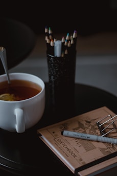 A warm and cozy setting features a white ceramic cup filled with tea, indicating a relaxed atmosphere. Colored pencils are neatly arranged in a black holder alongside an open sketchbook, suggesting artistic activity. A stylish pen rests on the sketchbook, ready for use.