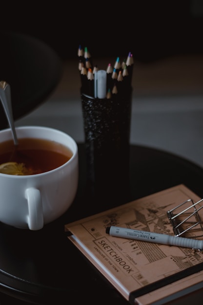 A cozy workspace with art supplies, sketchbooks, and a cup of tea beside a window filled with natural light.