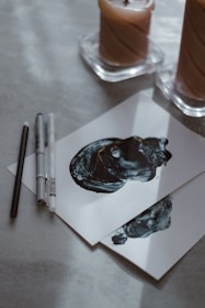 Two pieces of paper with abstract black ink artwork are placed on a light-colored surface. To the left of the papers, there are drawing tools, including a pencil and two pens. In the background, two candles in separate holders are partially in view.