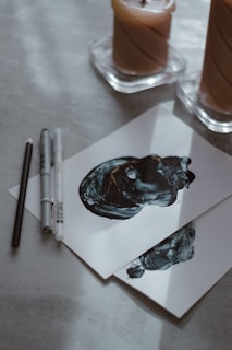 Two pieces of paper with abstract black ink artwork are placed on a light-colored surface. To the left of the papers, there are drawing tools, including a pencil and two pens. In the background, two candles in separate holders are partially in view.