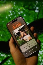 A close-up of a hand holding a smartphone displaying a music player interface with album art featuring two children. The background is blurred, showcasing lush green foliage.