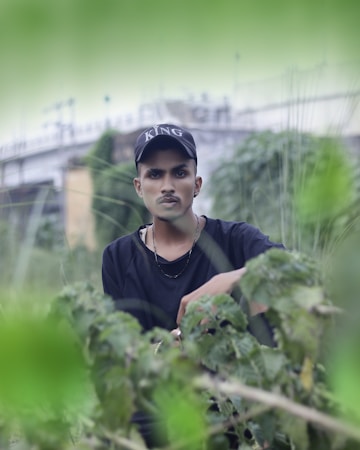 A young man wearing a black shirt and a cap with the word 'KING' on it stands amidst greenery. The background features blurred foliage and a distant bridge or architectural structure, creating a serene and natural atmosphere.