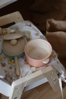 Two wooden toy cooking pots are placed on a piece of patterned fabric. One is an open pot with a pinkish hue, and the other has a lid with a pale blue rim. They rest on a light wooden structure that appears to be part of a child's play kitchen setup. A brown paper bag is visible in the background.