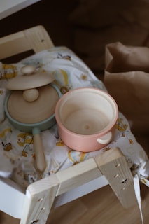 Two wooden toy cooking pots are placed on a piece of patterned fabric. One is an open pot with a pinkish hue, and the other has a lid with a pale blue rim. They rest on a light wooden structure that appears to be part of a child's play kitchen setup. A brown paper bag is visible in the background.