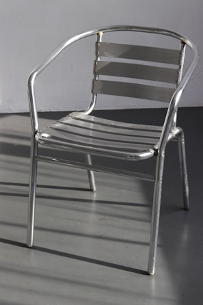 A metal chair with a sleek, minimalist design composed of horizontal slats and tubular metal frame. It casts shadows on a smooth gray floor, with a plain wall in the background.