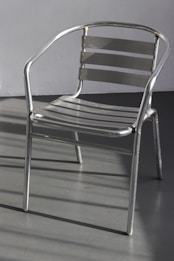 A metal chair with a sleek, minimalist design composed of horizontal slats and tubular metal frame. It casts shadows on a smooth gray floor, with a plain wall in the background.