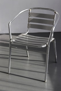 A metal chair with a sleek, minimalist design composed of horizontal slats and tubular metal frame. It casts shadows on a smooth gray floor, with a plain wall in the background.