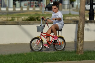 A young child with glasses is riding a bright red bicycle with a basket on the front. The child is wearing a white shirt with the word 'FRIENDS' printed on it, white shorts, and pink shoes. The setting appears to be a paved path in an outdoor area with grass in the foreground and a tree nearby.