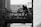 A monochrome image featuring a silhouette of a person watering plants on a small balcony or terrace. The background reveals rooftops and part of a building, suggesting an urban setting. Several potted plants are placed on the railing.