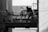 A monochrome image featuring a silhouette of a person watering plants on a small balcony or terrace. The background reveals rooftops and part of a building, suggesting an urban setting. Several potted plants are placed on the railing.