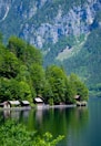Several small wooden cabins are nestled along the edge of a tranquil lake, surrounded by lush green trees. In the background, steep, rocky mountains rise dramatically, covered with patches of forest.
