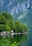 Several small wooden cabins are nestled along the edge of a tranquil lake, surrounded by lush green trees. In the background, steep, rocky mountains rise dramatically, covered with patches of forest.