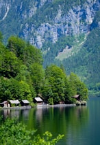 Several small wooden cabins are nestled along the edge of a tranquil lake, surrounded by lush green trees. In the background, steep, rocky mountains rise dramatically, covered with patches of forest.