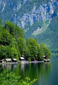 Several small wooden cabins are nestled along the edge of a tranquil lake, surrounded by lush green trees. In the background, steep, rocky mountains rise dramatically, covered with patches of forest.
