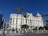 A grand, historic hotel with a pink dome and ornate architectural details is surrounded by palm trees. The clear blue sky creates a vivid backdrop, and a few vehicles are parked nearby. The large structure is meticulously designed, with multiple windows and decorative elements.